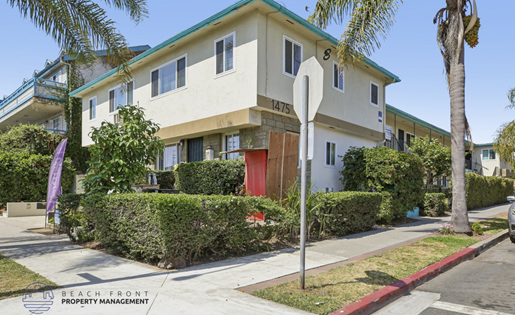 A white house with a green roof and a red door is surrounded by greenery and a palm tree.